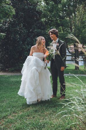 Happy bride and groom walking together on green lawn, bride in layered white gown holding colorful bouquet.