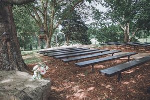 Rustic outdoor wedding ceremony setup with dark wooden benches under large trees and floral arch.