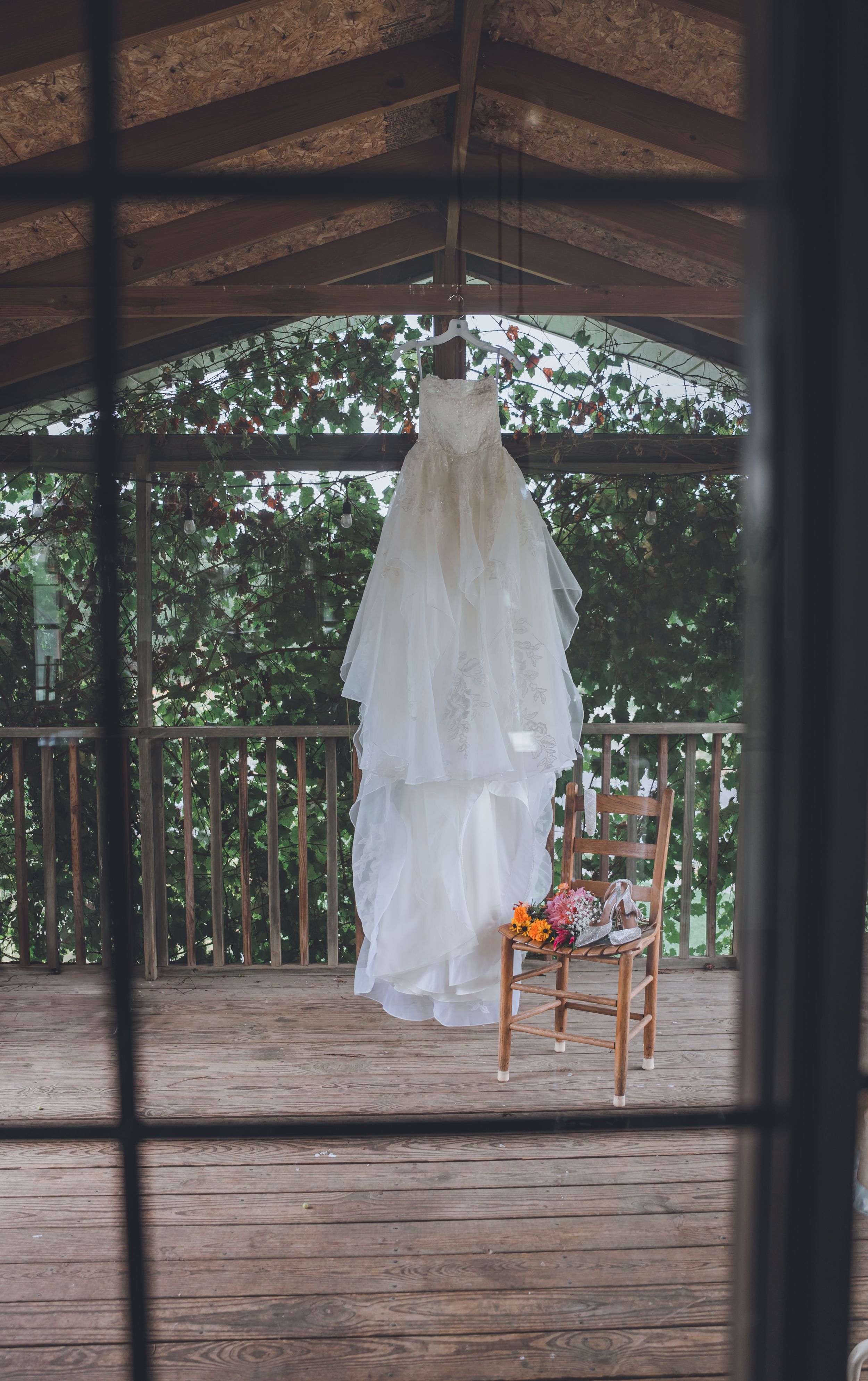 White wedding dress hanging under a rustic wooden gazebo with a chair below holding a colorful bridal bouquet.