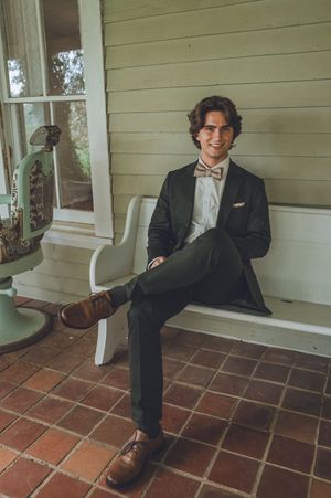 Dapper groom in black suit and bow tie sitting relaxed on white bench on rustic porch smiling.