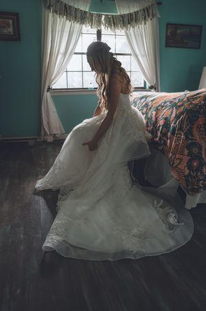 Bride in elegant white lace wedding gown preparing in a teal bedroom with hardwood floors and white curtains.