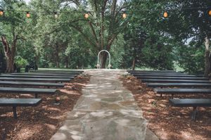 Outdoor wedding ceremony aisle lined with dark benches leading to a white arch surrounded by lush trees.