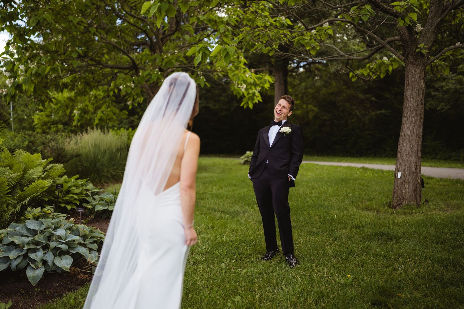 Bride in white gown and veil faces groom in tuxedo during outdoor first look ceremony in lush garden.