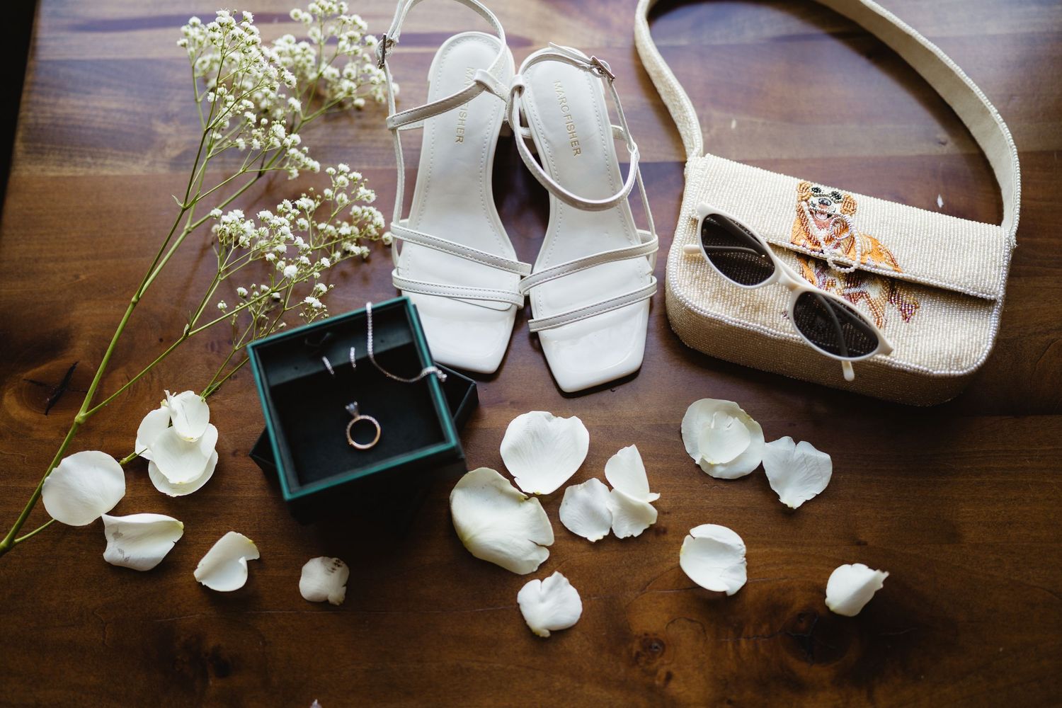 Flat lay of white strappy sandals, ring box, floral purse, sunglasses, and rose petals on wooden surface.
