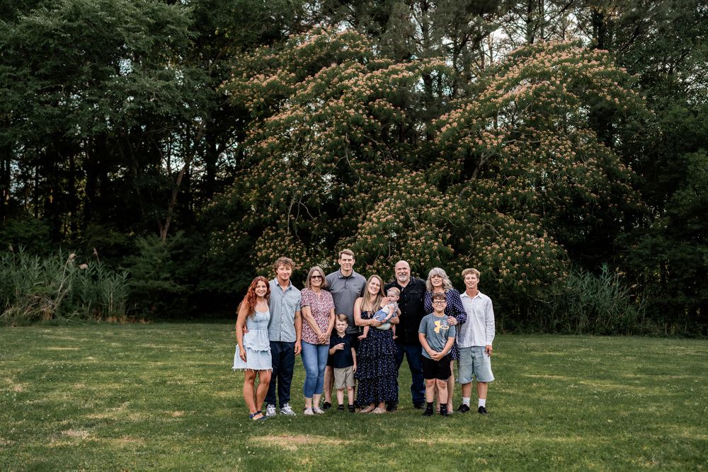 Large family group portrait outdoors on green lawn with lush trees in background on a summer day.