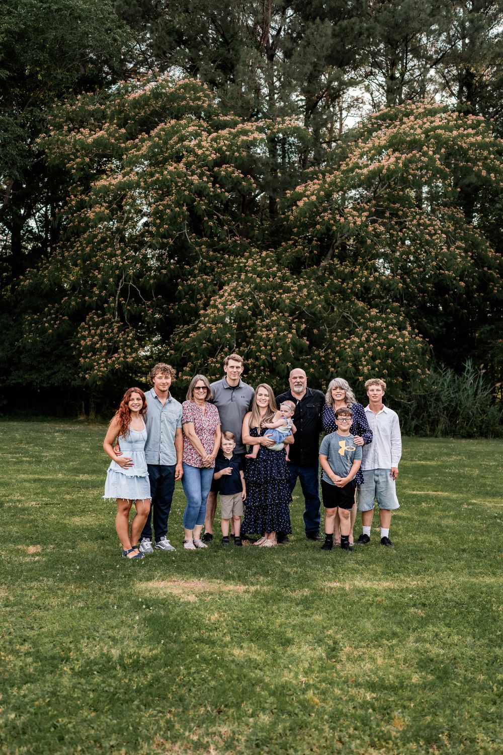 Large multigenerational family posing together on green lawn with blooming tree backdrop during outdoor photo session.