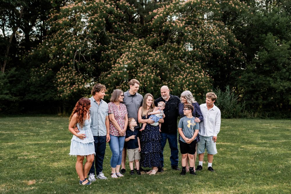 Large family group portrait outdoors on green lawn with lush trees, including children and adults smiling together.