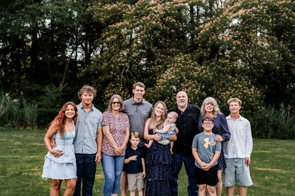 Extended family group of ten smiling together on grass with lush green trees in background.