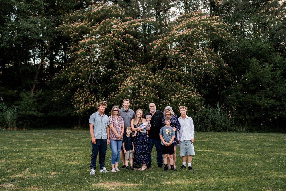 Family of nine posing outdoors on green lawn with blooming white tree backdrop in warm evening light.