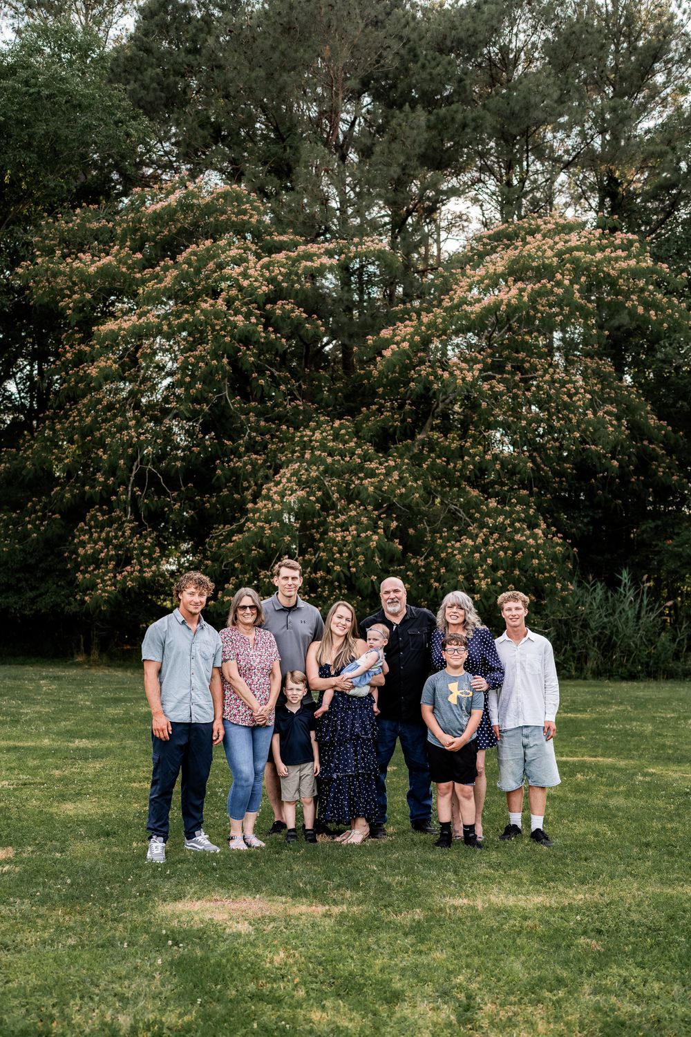 Multi-generational family group portrait outdoors on green lawn with blooming tree backdrop at golden hour.