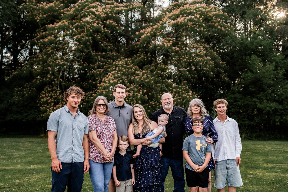 Large multigenerational family posing together outdoors on green grass with lush trees at golden hour.