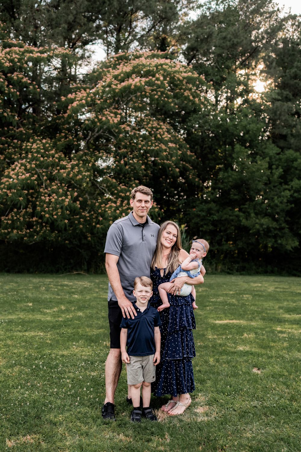 Happy family of four posing together on green grass with blooming trees behind them.