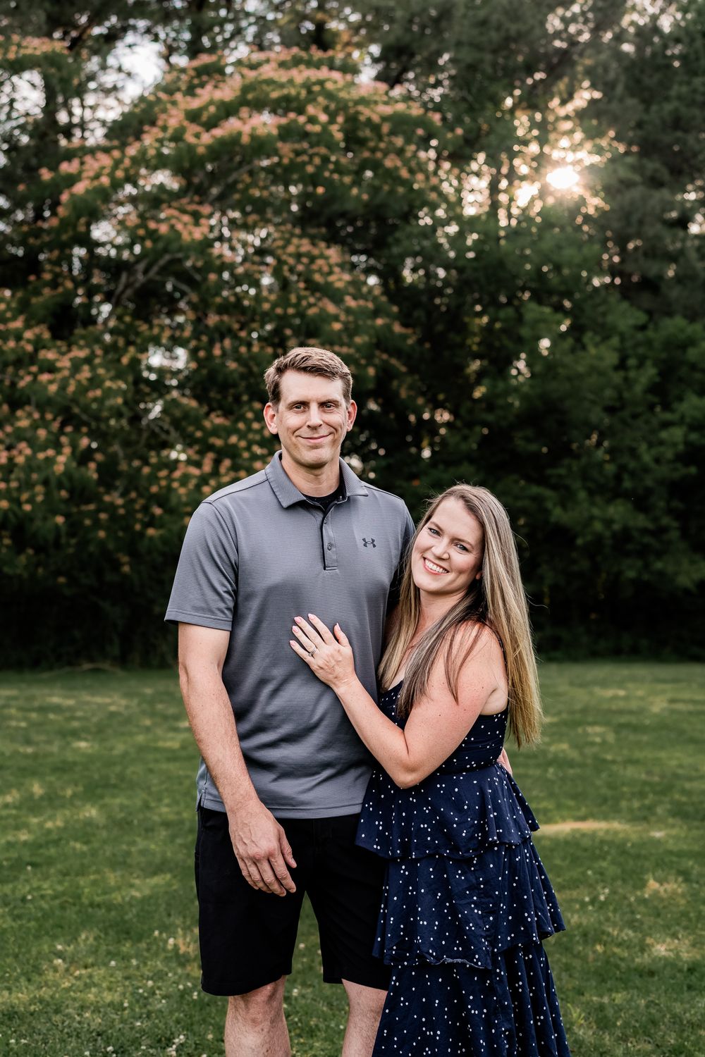 Smiling engaged couple posing in lush green park at sunset, woman showing ring.