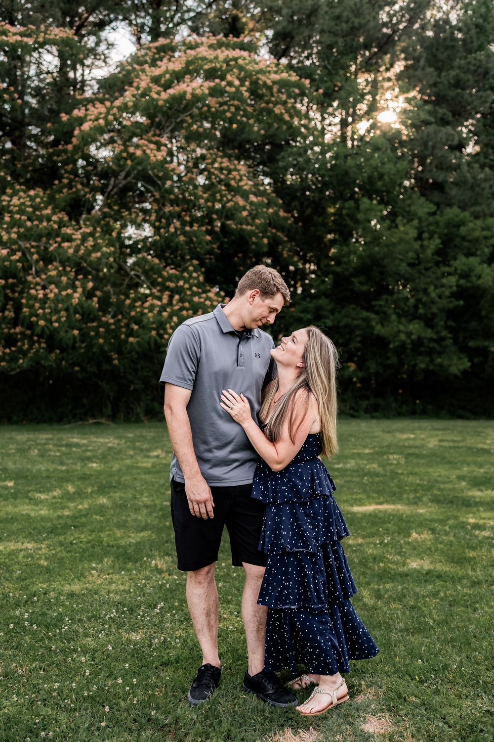Couple sharing a romantic moment in a lush green park, woman in navy dress, man in gray polo, blooming trees behind.