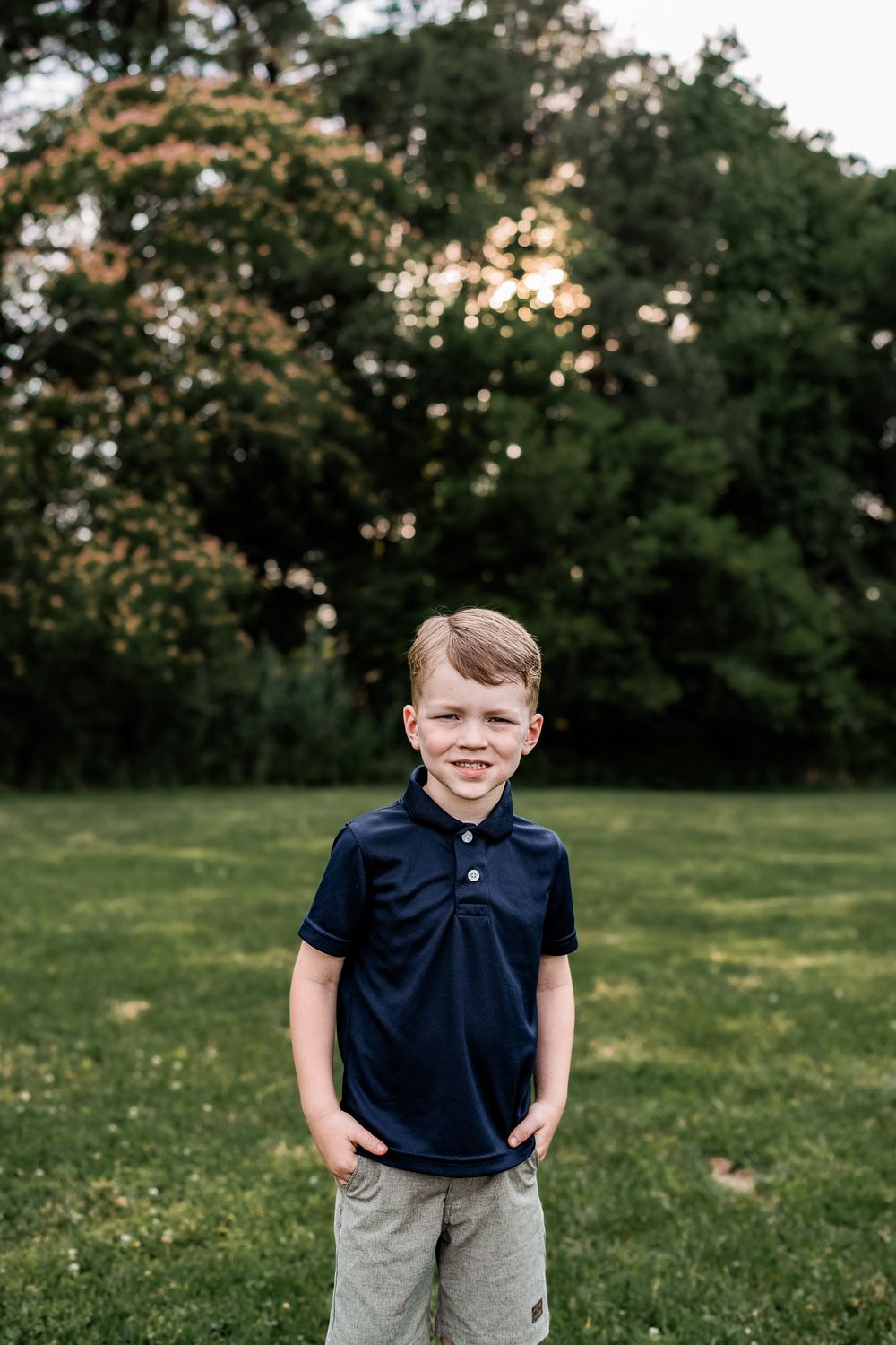 Smiling young boy in a navy polo shirt standing on green grass with trees in the background.