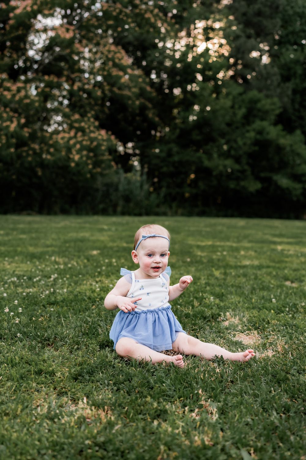 Happy baby girl in blue romper sitting on green grass outdoors at sunset.