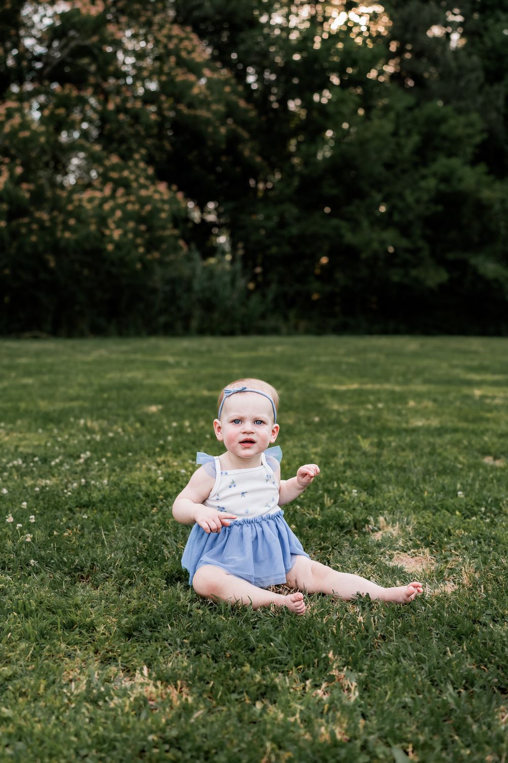 Adorable baby girl in blue romper sitting on green grass at dusk in a park.