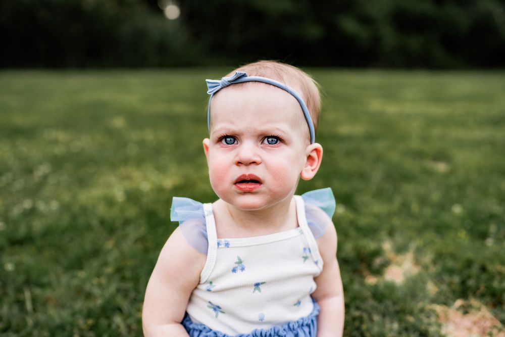 Adorable baby girl with a blue bow headband sitting on grass with a curious expression.