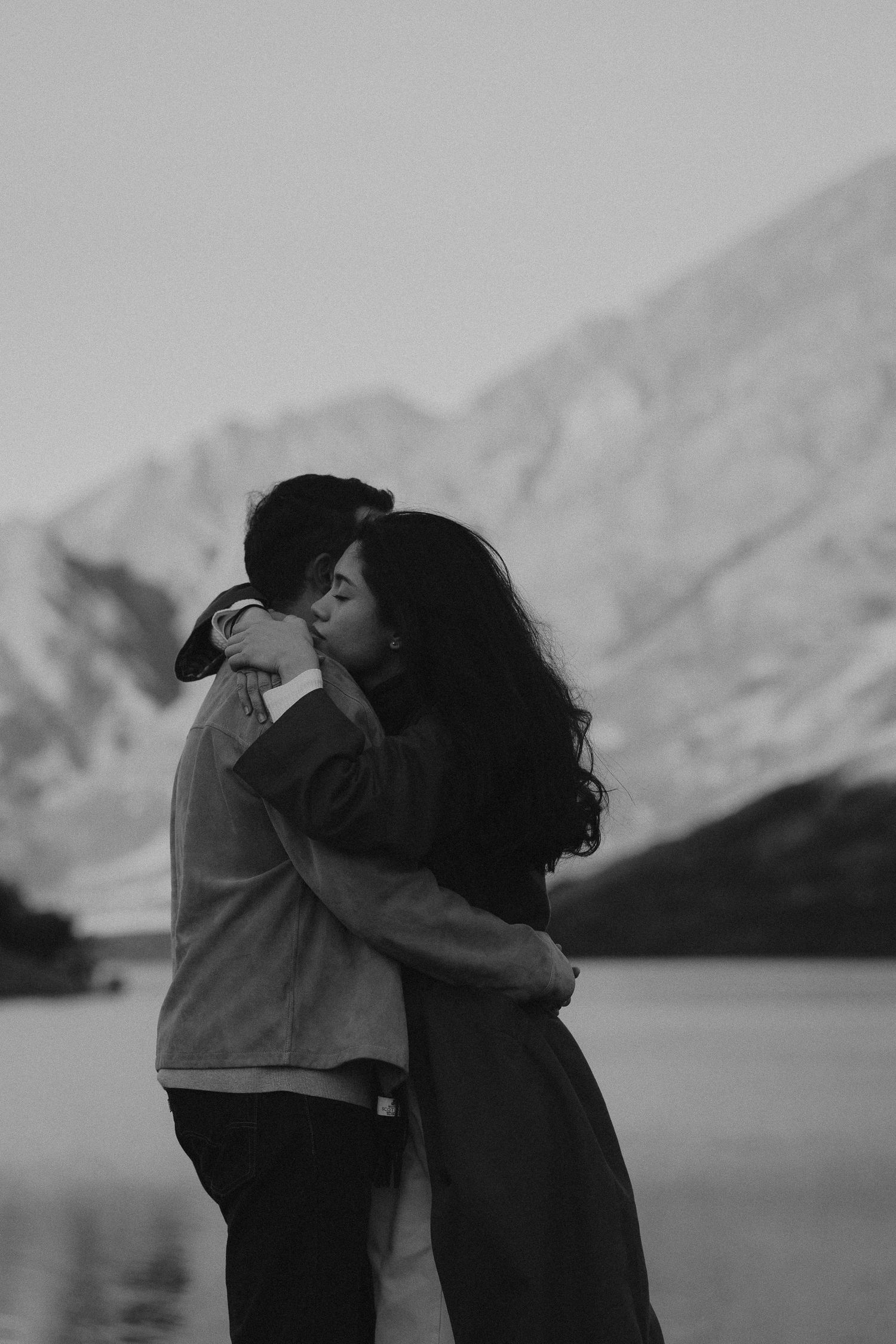 Couple embracing by a serene mountain lake at golden hour, with dramatic rocky peaks glowing in warm sunlight behind them.