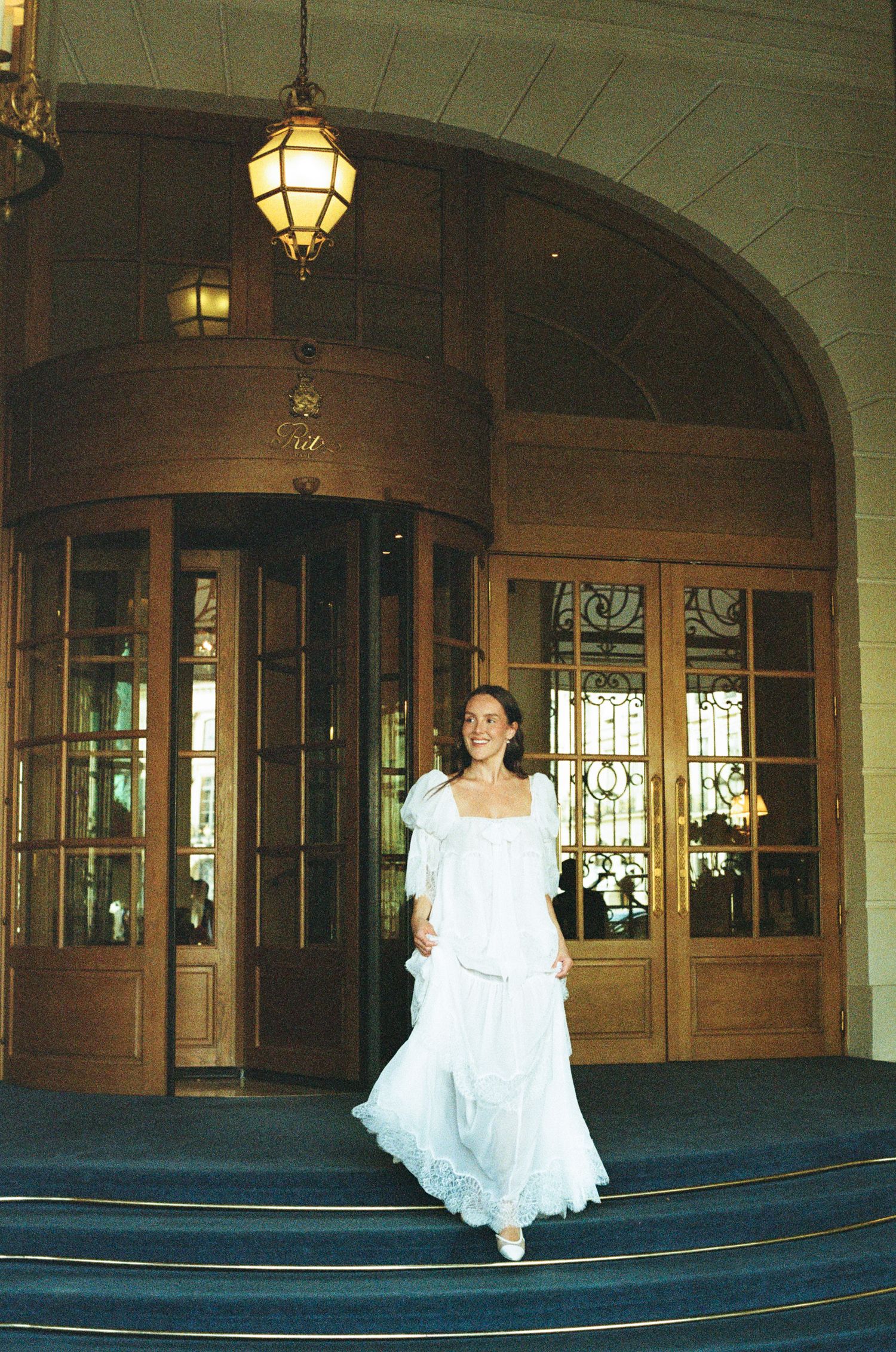 A woman in a white flowing dress walks down steps outside the elegant Ritz Hotel entrance with ornate wooden doors.