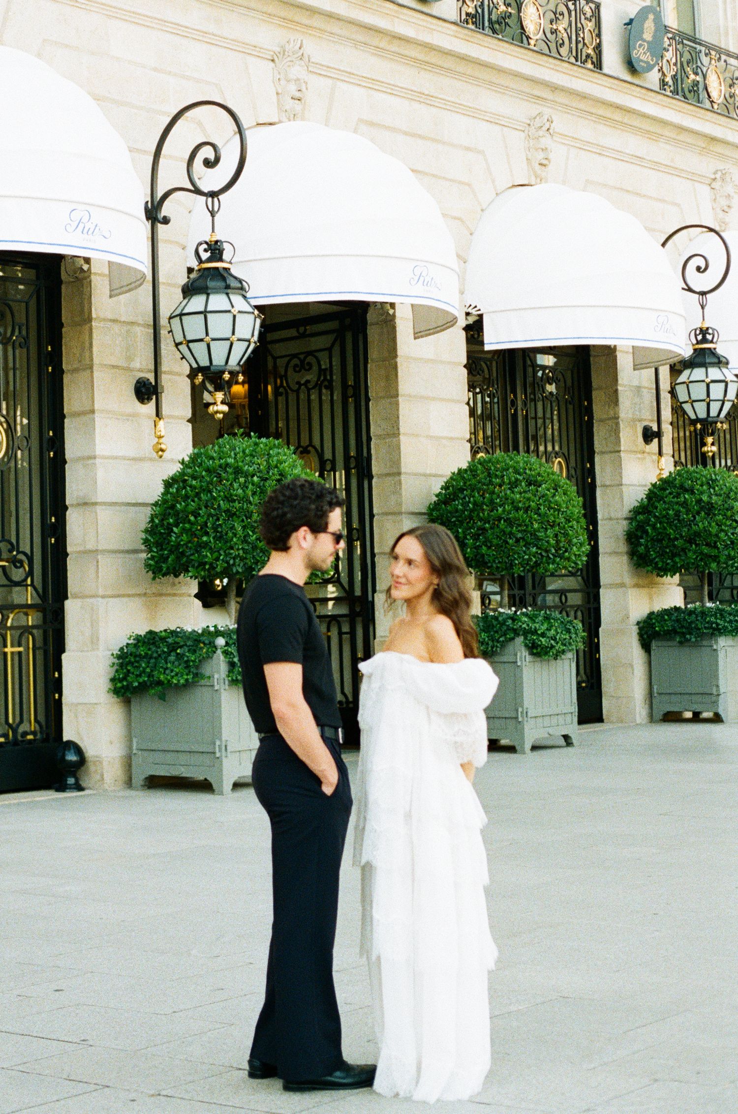 A couple in elegant attire stands facing each other outside a Parisian hotel with white awnings and lush greenery.