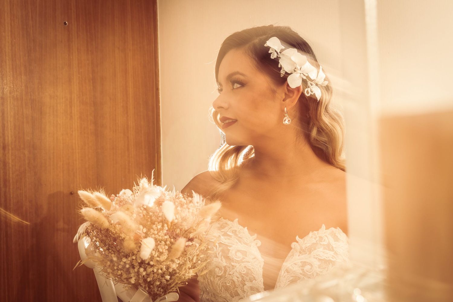 Bride holding dried floral bouquet wearing lace gown and floral hair accessory, bathed in warm golden light.