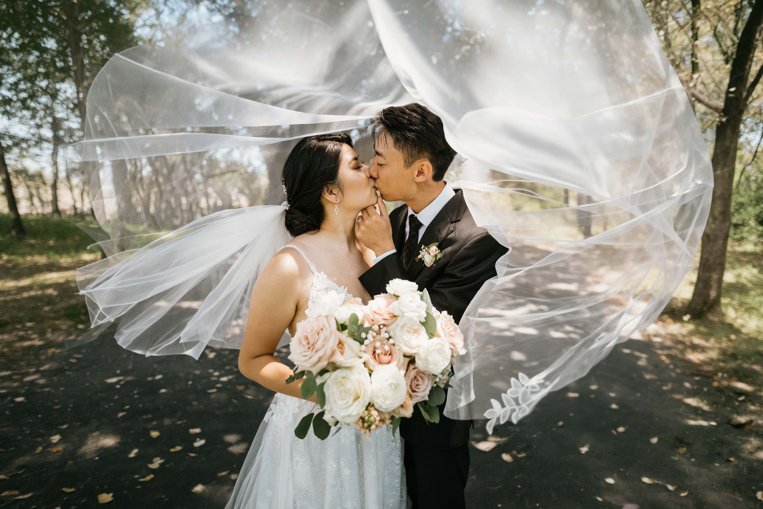 Bride and groom sharing a kiss beneath the bride's flowing veil during romantic wedding portraits on a tree-lined path.