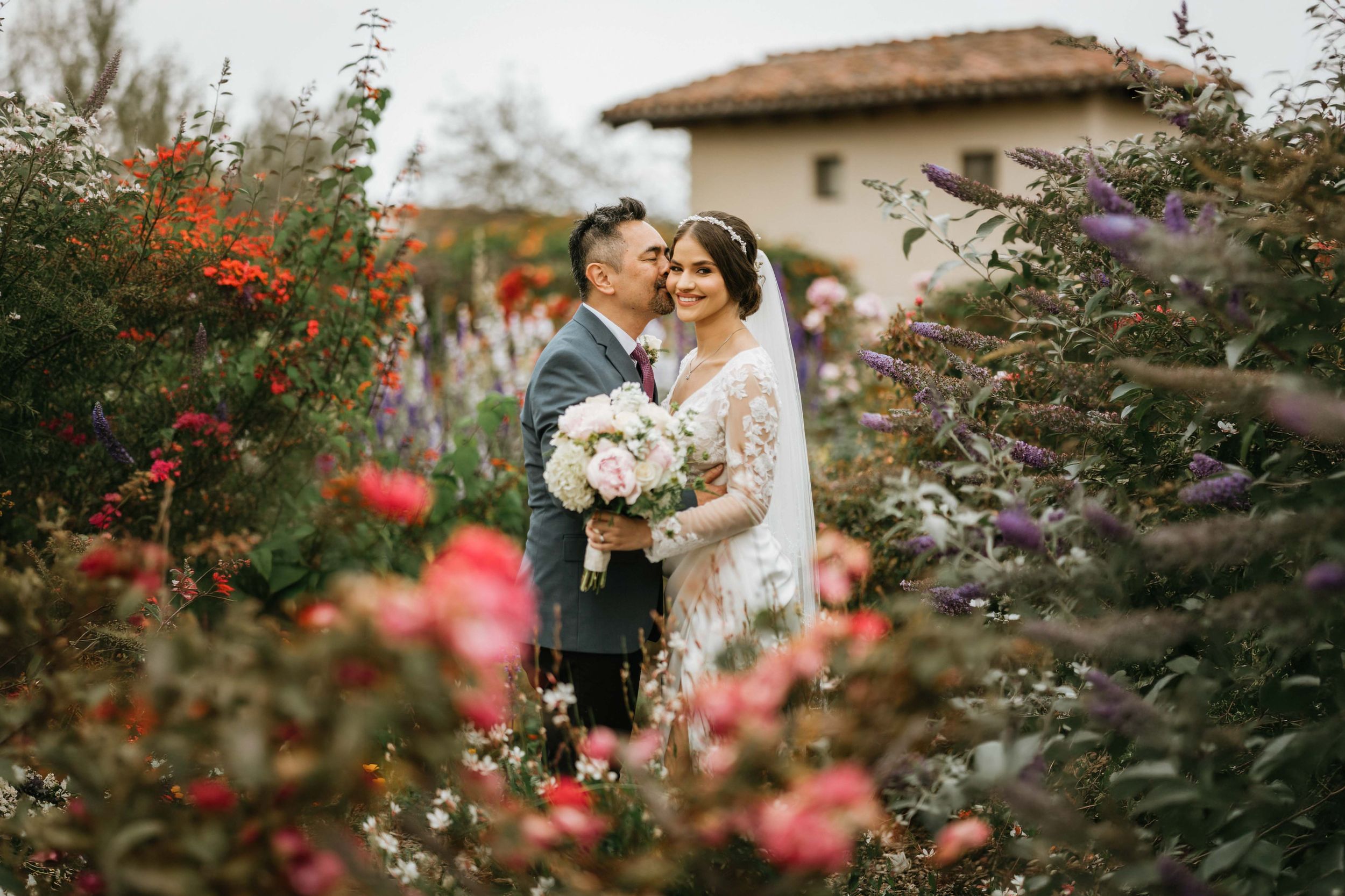 Bride and groom sharing a romantic portrait surrounded by vibrant garden flowers at Mission San Juan Capistrano in San Juan Capistrano, Cali