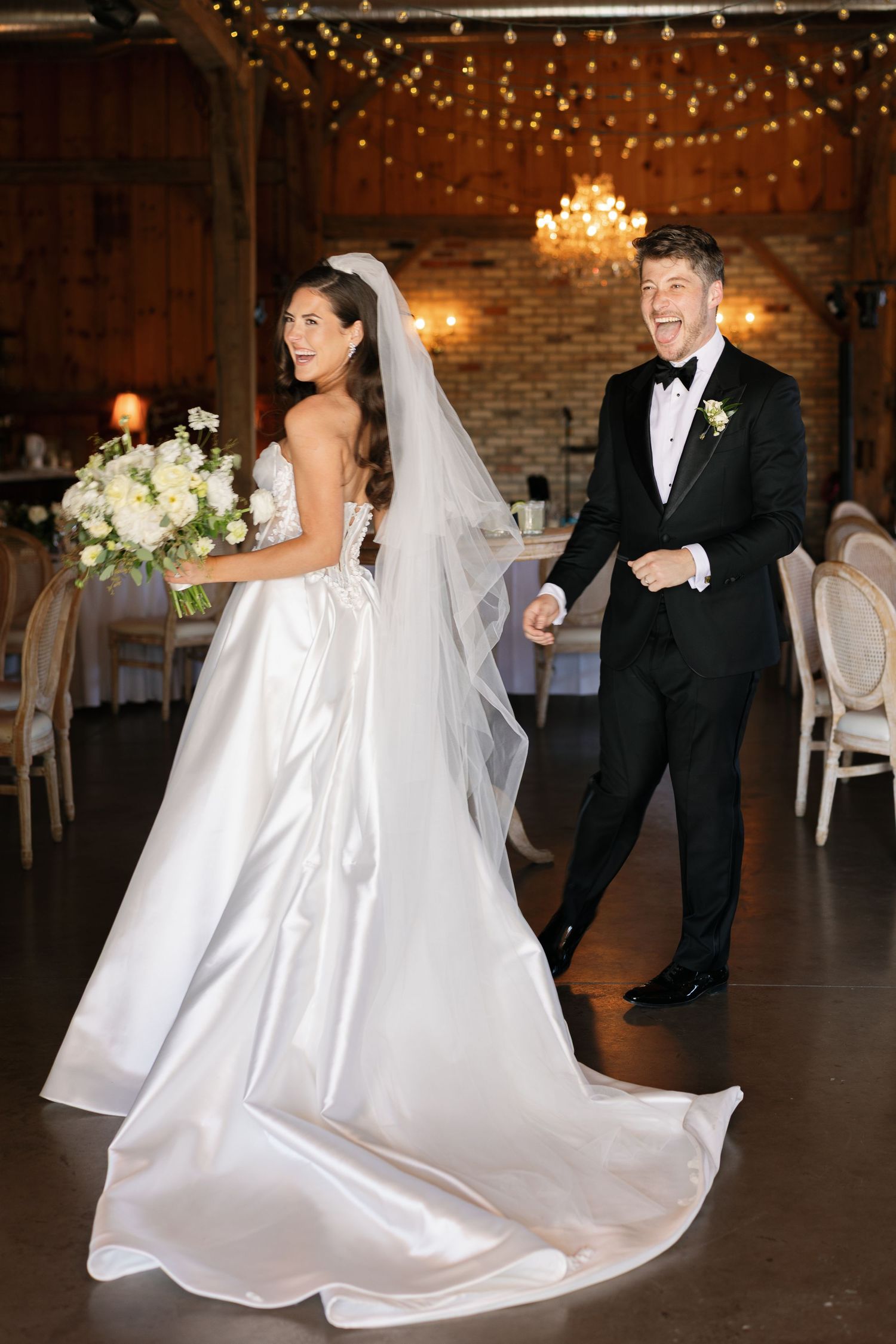 Bride in white satin gown with veil holds bouquet as groom in black tuxedo laughs during wedding reception.