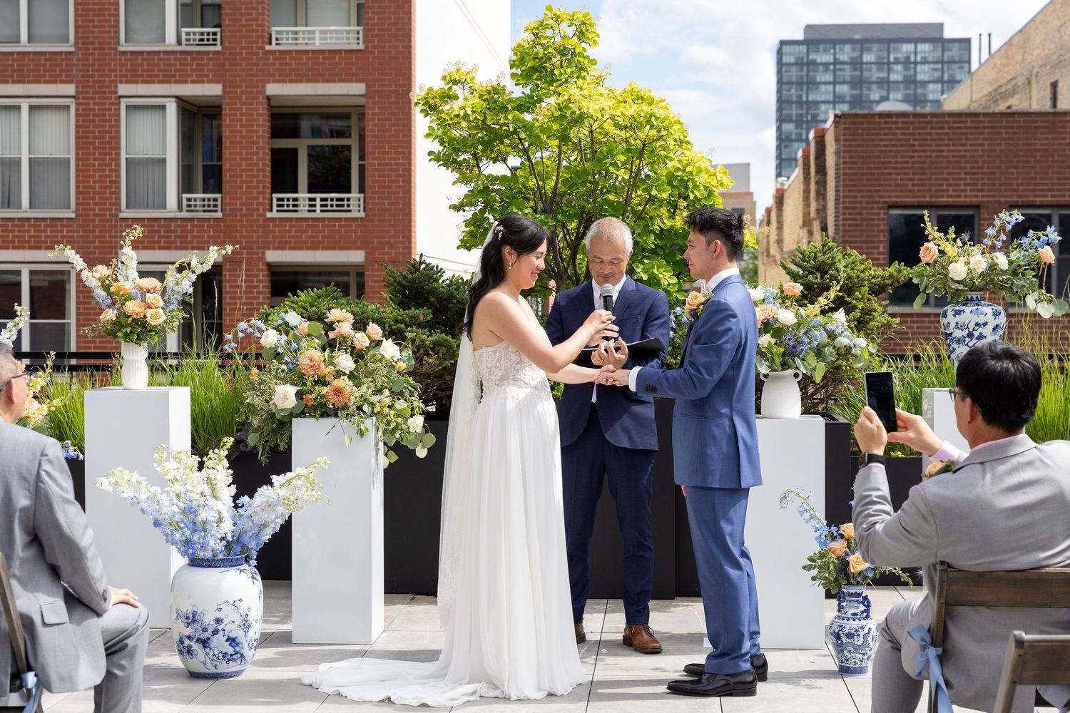 Couple exchanges vows at outdoor rooftop wedding ceremony with floral arrangements and city buildings in background.