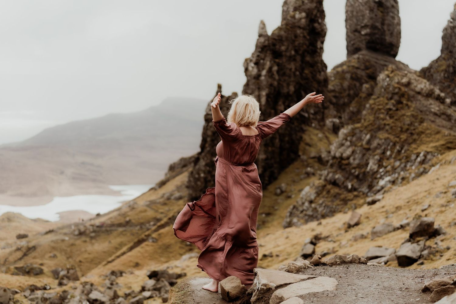 Woman in flowing burgundy dress with arms raised at the Old Man of Storr, Isle of Skye, Scotland.