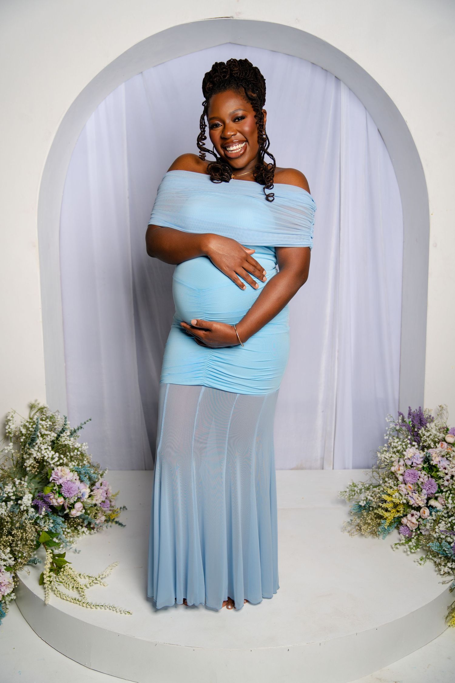 Smiling pregnant woman in light blue off-shoulder gown poses in front of arch backdrop with floral arrangements.