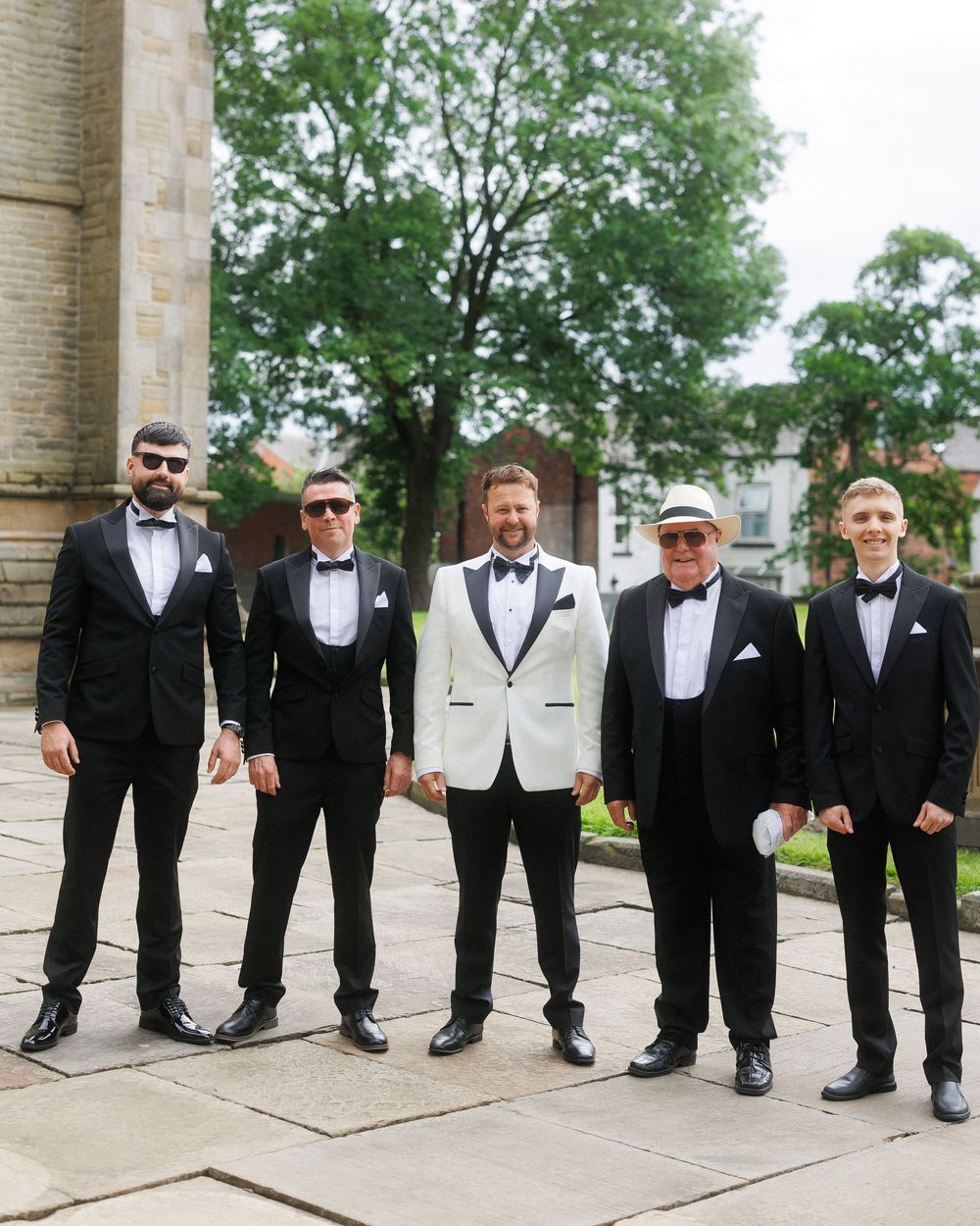 Five men in black tuxedos and bow ties pose outdoors, with one wearing a white jacket and another in a white hat.