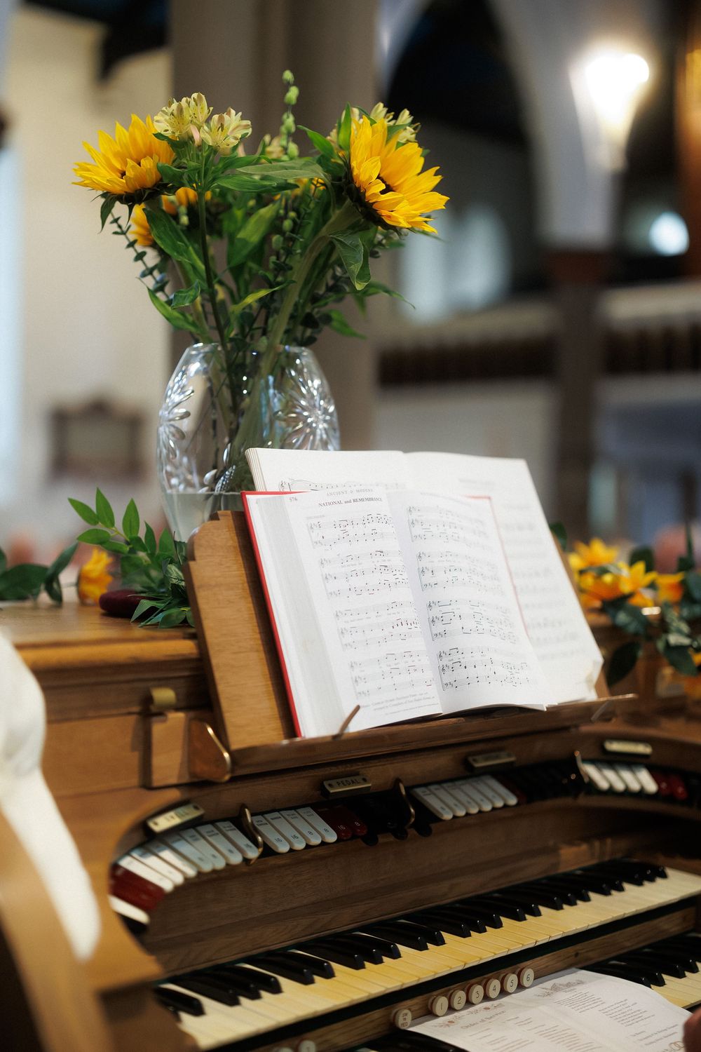 Wooden church organ with open sheet music book on stand and yellow sunflowers in glass vase, elegant musical setting.