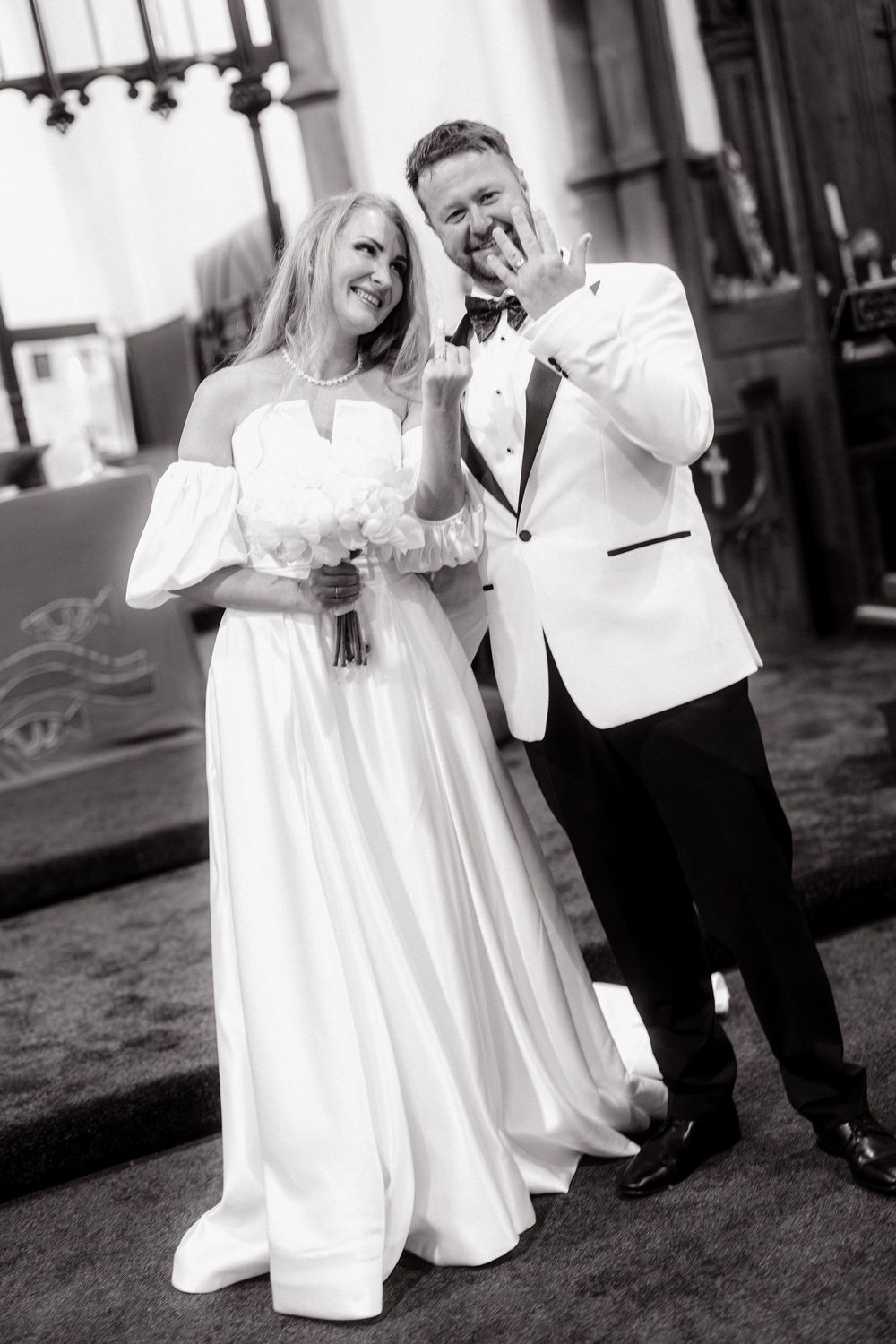 Black and white photo of smiling bride holding bouquet and groom in white tuxedo jacket posing together at wedding.