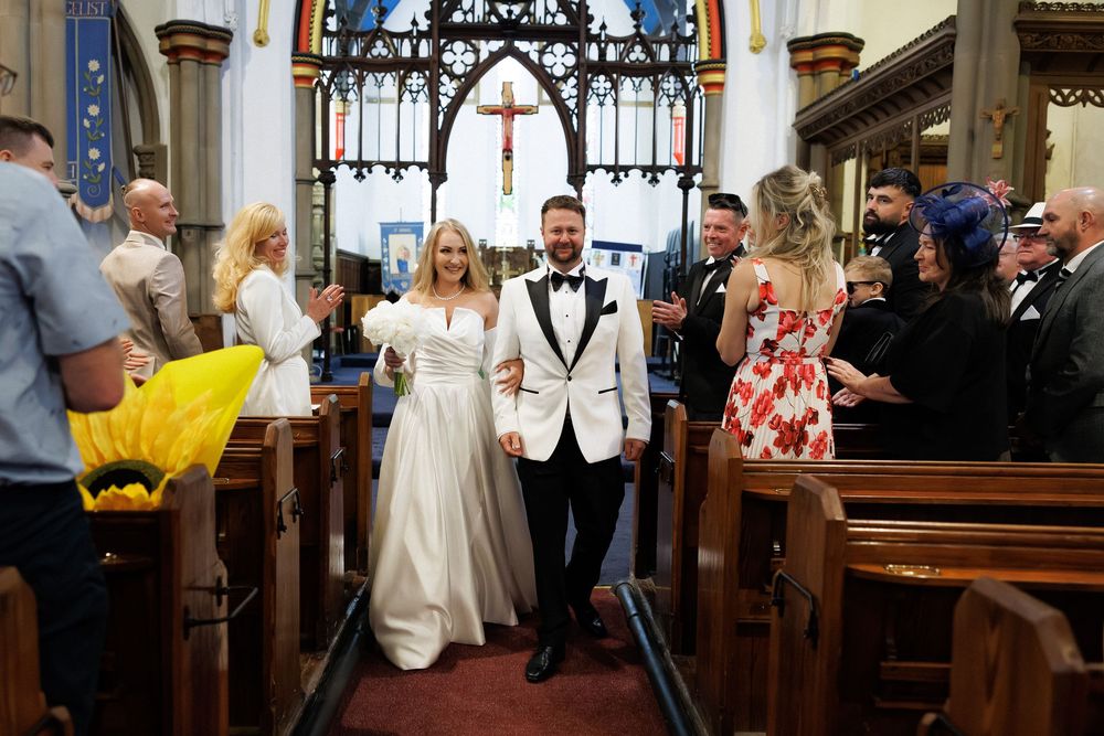 Joyful newlyweds walk down church aisle after wedding ceremony, bride in white gown, groom in tuxedo, guests cheering.
