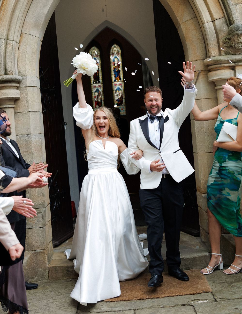 Joyful newlywed couple exits church in wedding attire as guests toss confetti petals in celebration.