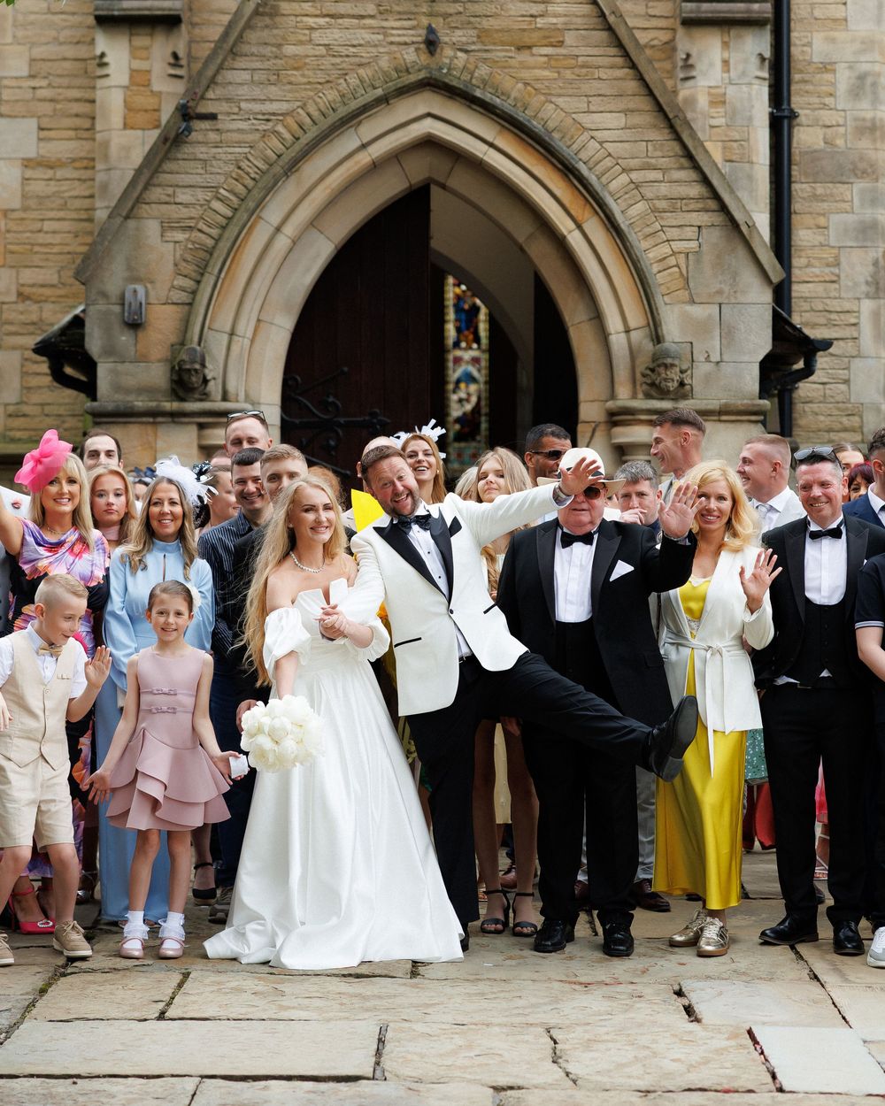 Joyful wedding party celebrating outside a stone church, bride in white gown holding bouquet, groom in tuxedo, guests cheering.