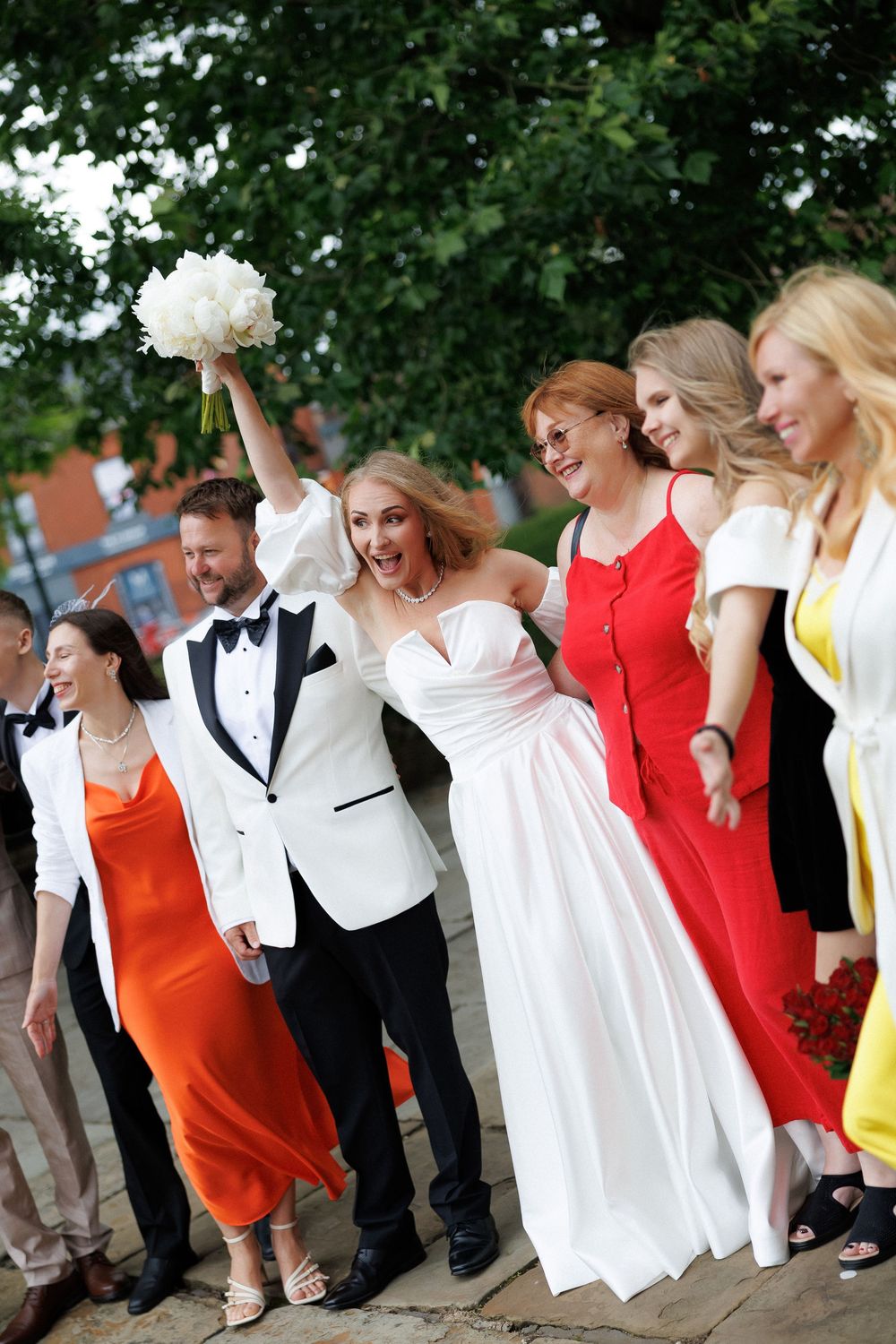 Joyful bride and groom celebrate with wedding party outdoors, bride raising bouquet of white flowers.