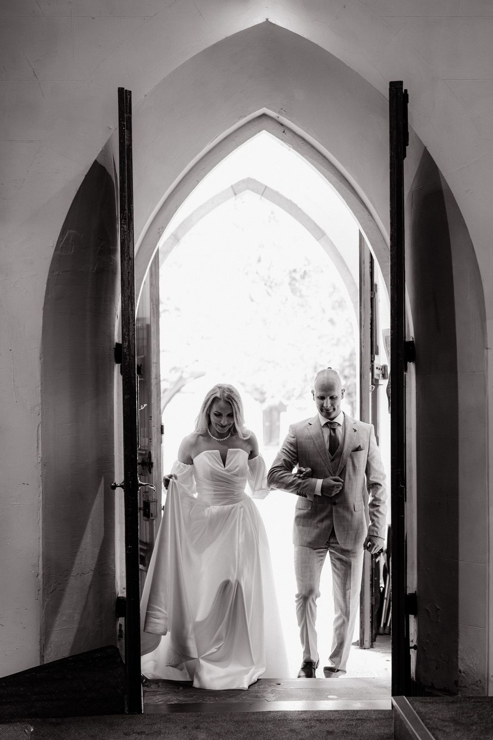 Black and white photo of bride and groom entering church through gothic arched doors on wedding day.