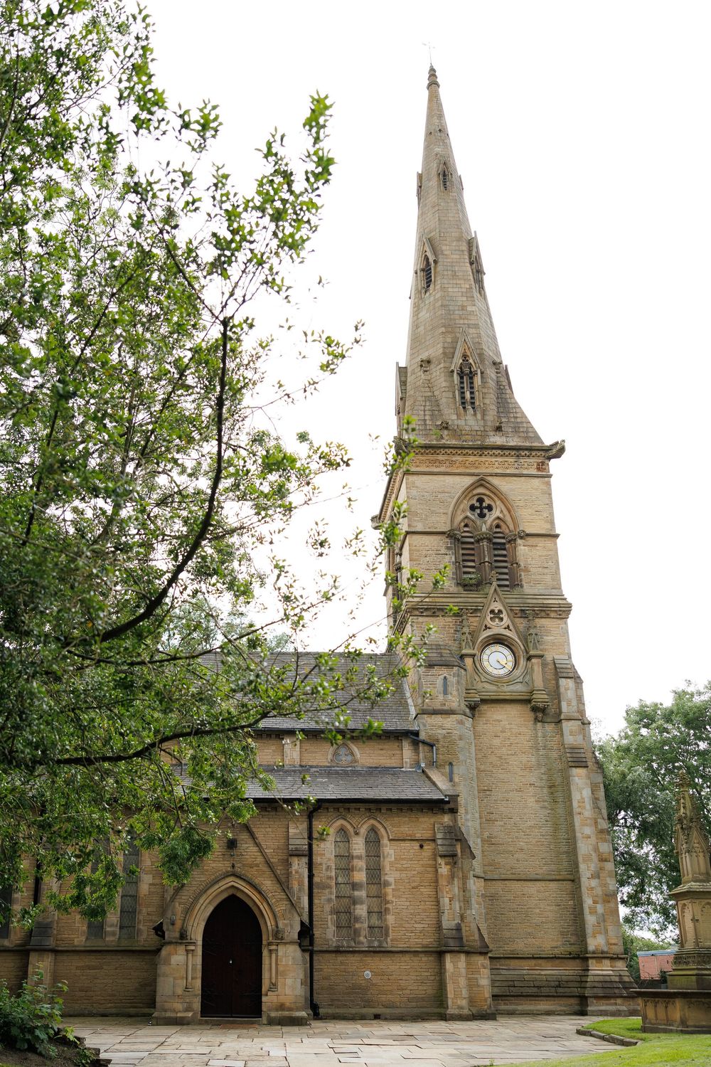 Historic Gothic stone church with tall spire and arched entrance, surrounded by trees under overcast sky.