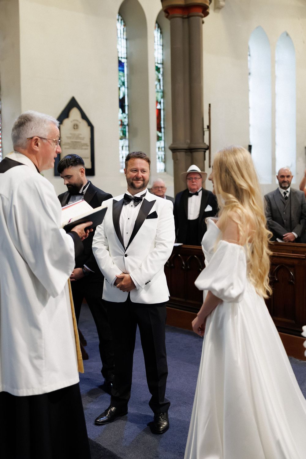 Groom in white tuxedo smiling at bride in off-shoulder gown during church wedding ceremony.