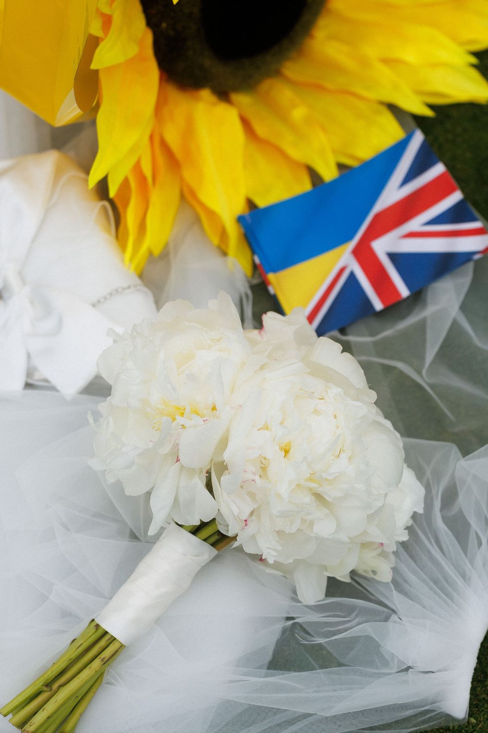 White peony bridal bouquet with Ukrainian and UK flags beside sunflowers on white tulle veil.