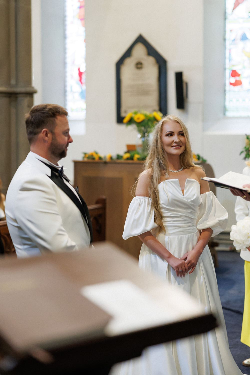 Bride in off-shoulder white gown and groom in white tuxedo exchange vows at a sunlit church altar.