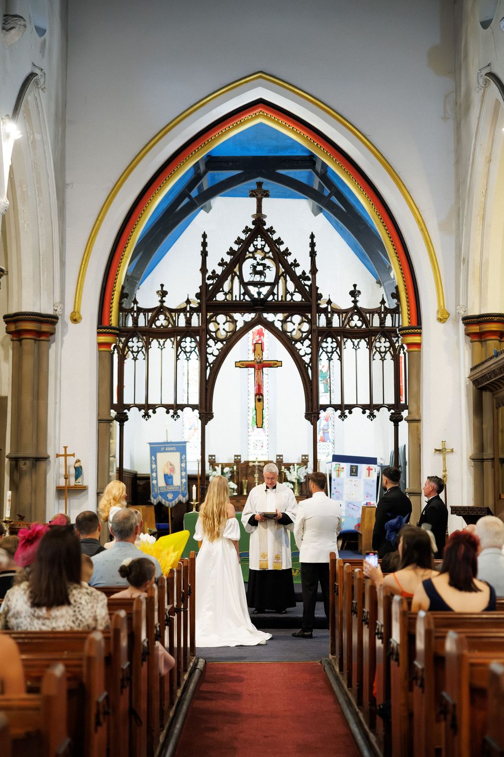 A bride and groom stand at the altar of a colorful Gothic church during their wedding ceremony with guests seated in pews.