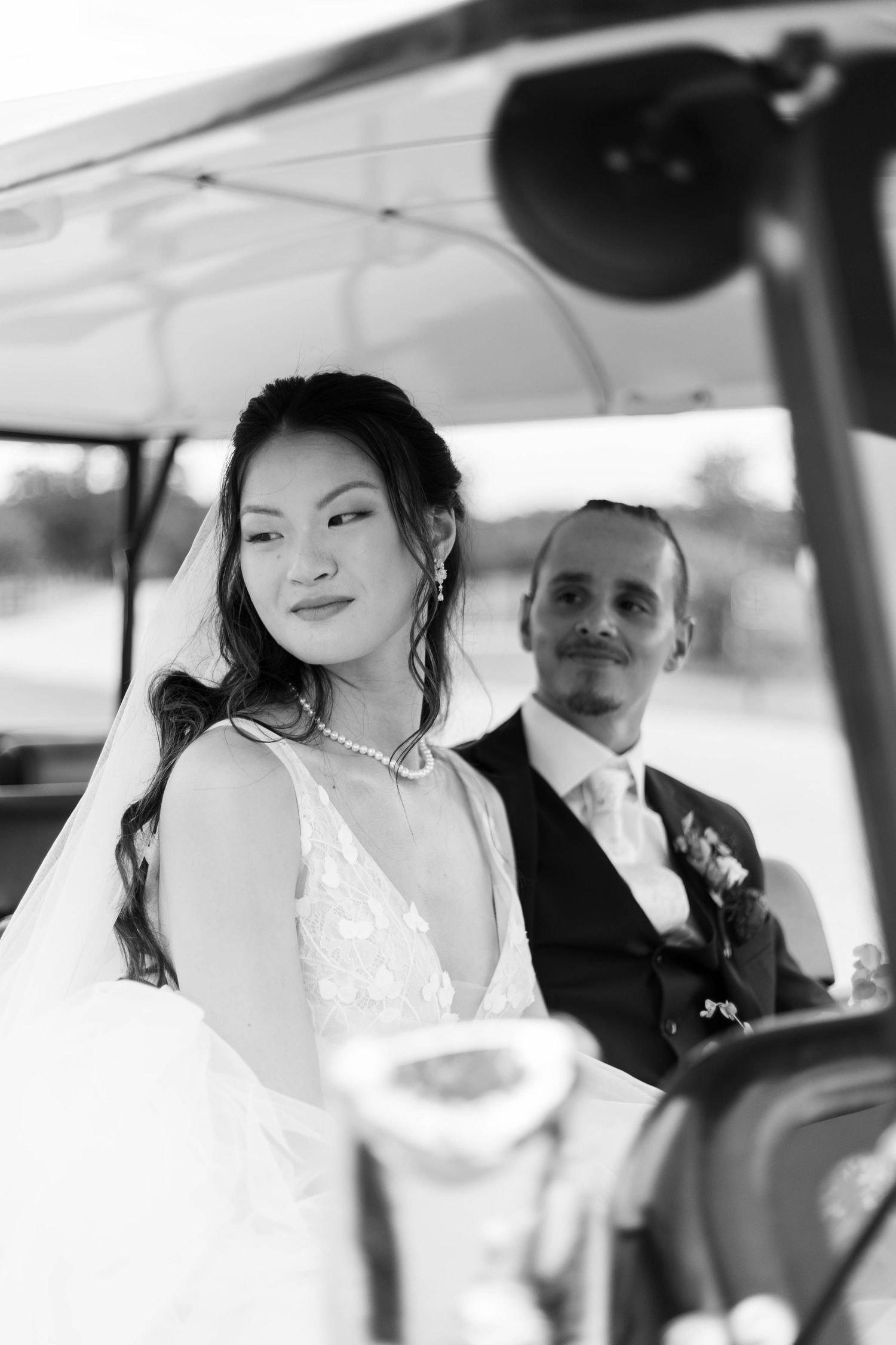 Black and white photo of a bride and groom riding in a golf cart on their wedding day.