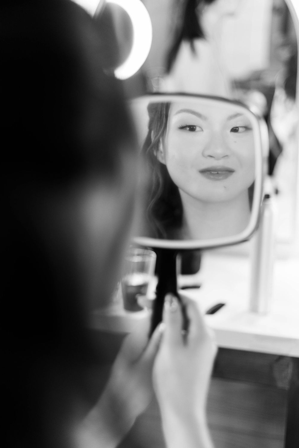 Black and white photo of a woman applying makeup while viewing her reflection in a lighted vanity mirror in Hummingbird Hill Wedding Venue