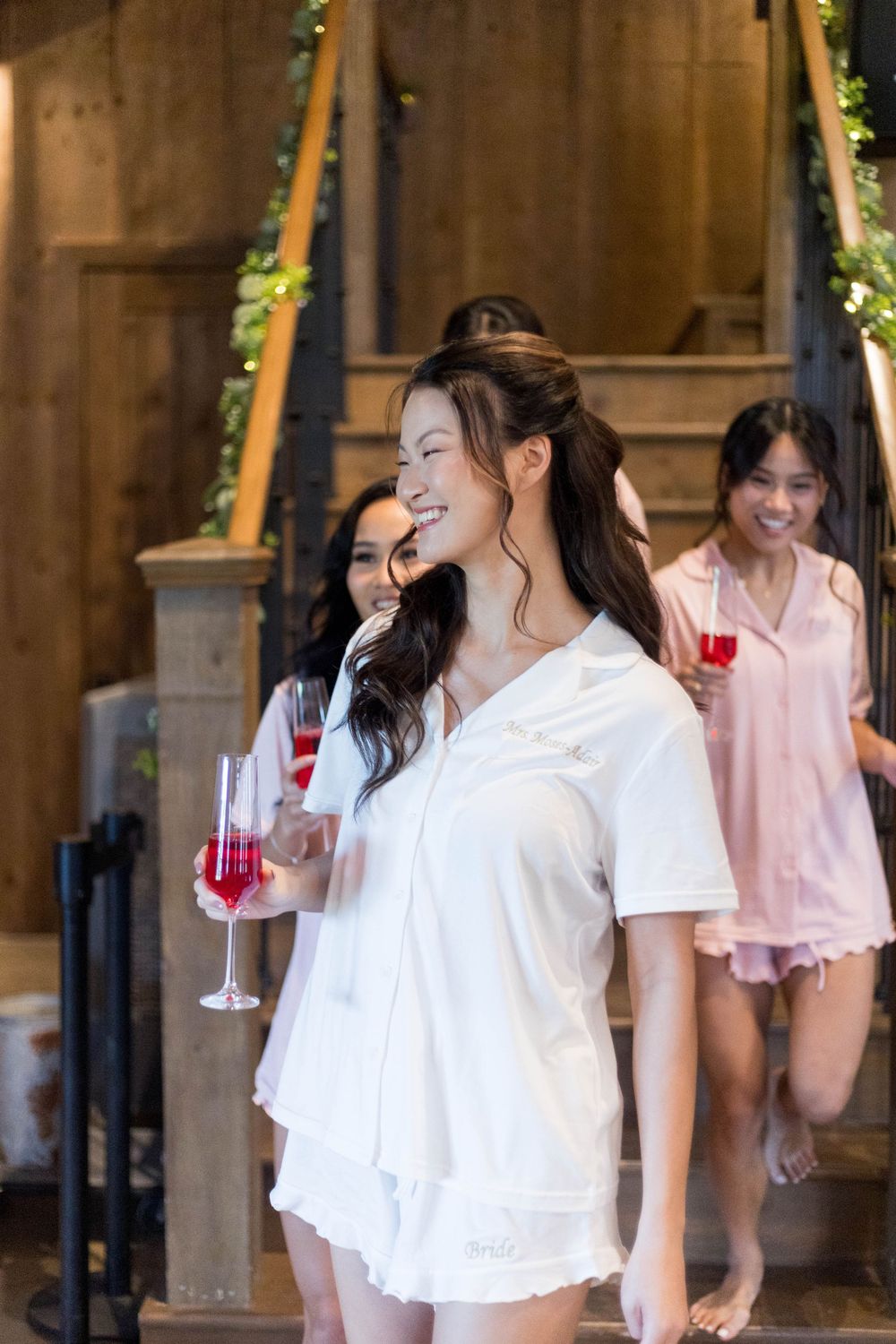Smiling bride in white pajamas holds champagne flute with bridesmaids descending rustic wooden staircase.