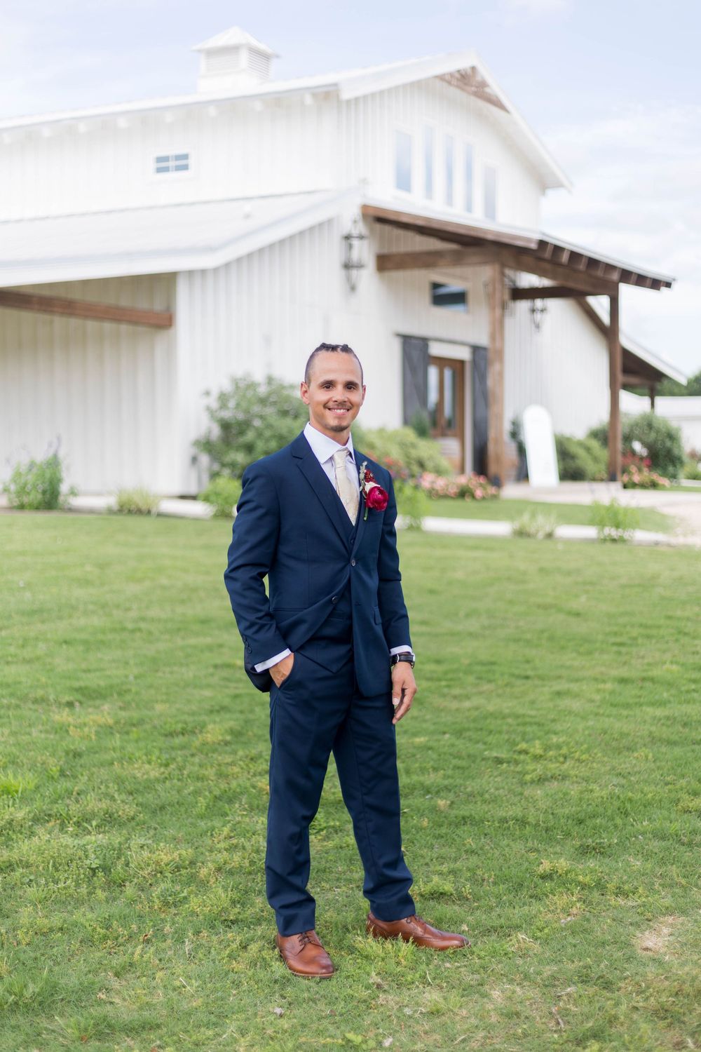 Groom in navy blue suit with boutonniere stands on green lawn before white barn wedding venue.