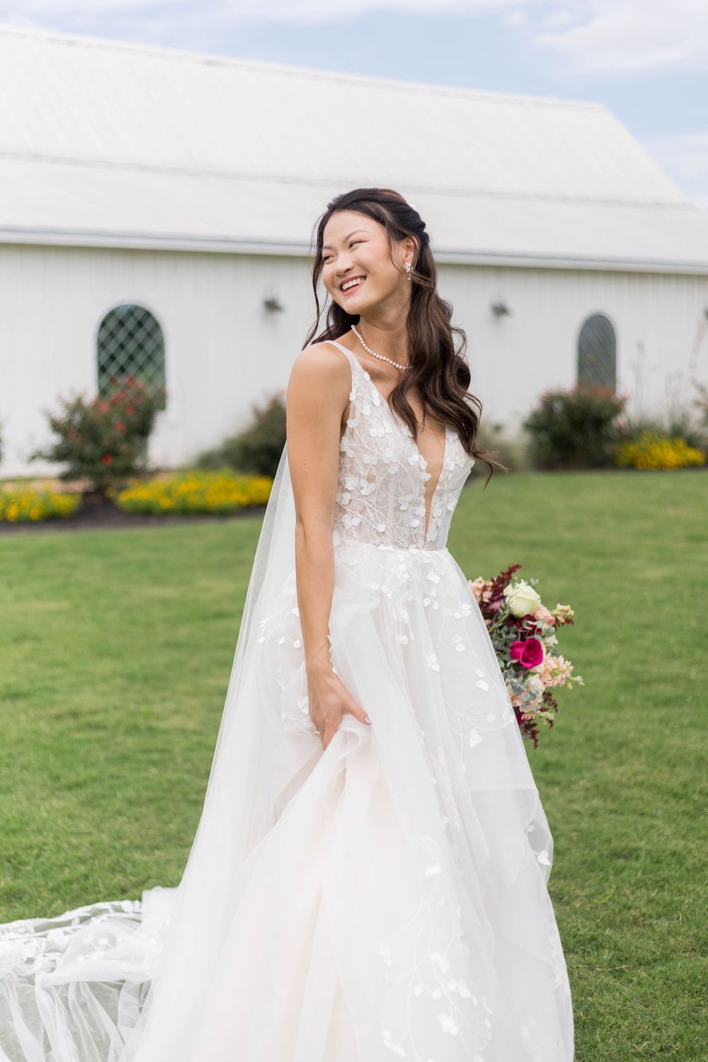 Smiling bride in floral lace ballgown holding bouquet on green lawn before white wedding venue at Hummingbird Hill